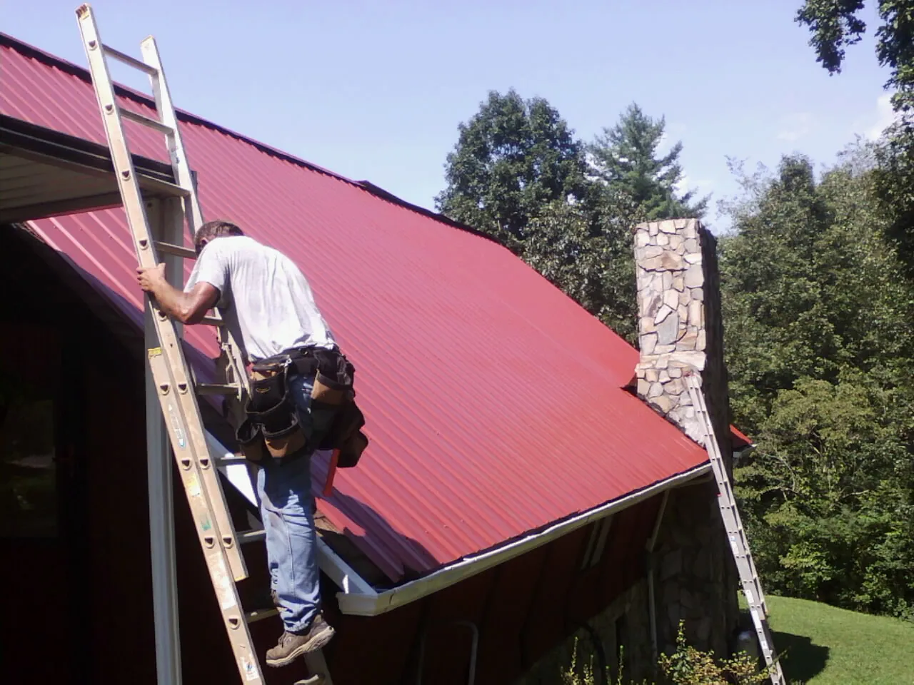 Roofer on a ladder beside a red metal roof and stone chimney — Soffit & Fascia Repair by Roofing Brothers in Johnson City